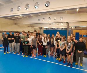 Grupa uczniów stojąca w hali sportowej, przygotowana do zajęć ruchowych; w tle elementy infrastruktury sportowej.