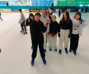 Uczennice na lodowisku podczas nauki jazdy na łyżwach; w tle inni uczestnicy zajęć.