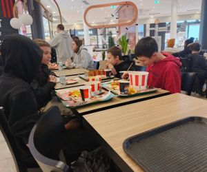 Uczniowie siedzący przy stolikach w strefie gastronomicznej centrum handlowego podczas przerwy obiadowej.