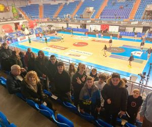 Uczniowie na trybunach hali widowiskowo-sportowej podczas meczu koszykówki; w tle boisko i zawodnicy rozgrzewający się przed spotkaniem.