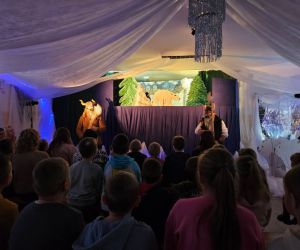 Dzieci siedzące na ławkach przed sceną, oczekujące na rozpoczęcie przedstawienia w sali teatralnej.