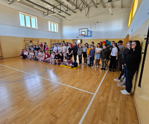 Uczniowie oraz dzieci z oddziałów przedszkolnych stoją i siedzą wzdłuż ściany sali gimnastycznej, obserwując program przygotowany z okazji Dnia Kobiet.