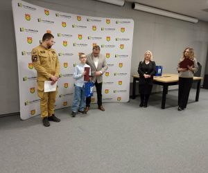 Moment wręczenia nagrody Aronowi Puchale przez przedstawiciela władz gminy. Obok stoi strażak oraz organizatorzy konkursu.