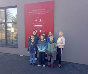 Grupa uczniów wraz z opiekunem stoi przed budynkiem Centrum Dziedzictwa Archeologicznego i Kulturowego w Masłomęczu.