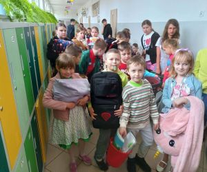 Uczniowie ustawieni na korytarzu trzymają różnorodne przedmioty użyte zamiast plecaków, pokazując swoją kreatywność.