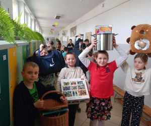 Uczniowie na szkolnym korytarzu prezentują nietypowe „plecaki” w ramach Dnia bez plecaka – wśród nich znajdują się m.in. garnek, koszyk i torby.