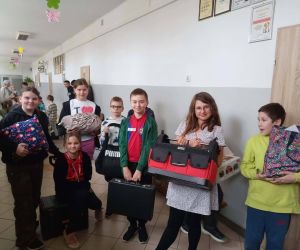 Grupa uczniów prezentuje swoje nietypowe „plecaki” na szkolnym korytarzu, trzymając różne przedmioty zastępujące tornistry.