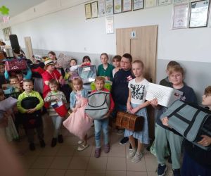 Uczniowie na korytarzu prezentują swoje rzeczy przyniesione zamiast plecaków, trzymając m.in. poduszki, wiadra i torby.