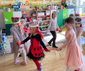 Dzieci w sali przedszkolnej biorą udział w zabawie ruchowej, trzymając się za ręce i poruszając się w kole.