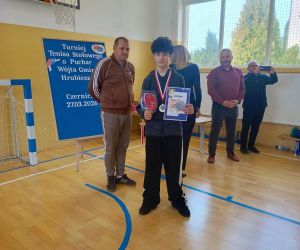 Uczeń z medalem i dyplomem po zakończeniu turnieju, w tle organizatorzy i tablica informacyjna.