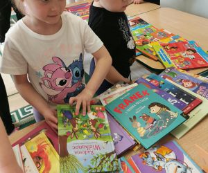 Dzieci stoją przy stoliku i przeglądają różnorodne książki dla dzieci, wybierając interesujące je tytuły.