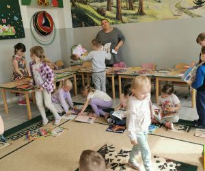 Dzieci w sali przedszkolnej wybierają książki ze stołu, a nauczyciel nadzoruje przebieg zajęć.