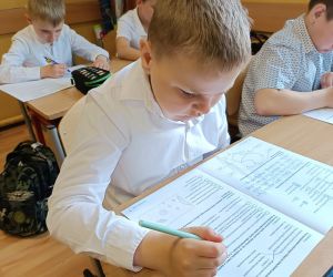 Uczeń w białej koszulce pracuje nad arkuszem sprawdzianu przy szkolnej ławce.