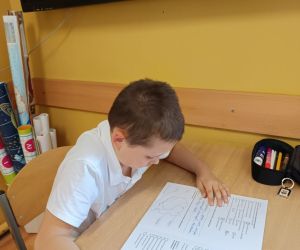 Uczeń siedzi przy ławce i rozwiązuje arkusz sprawdzianu, skupiony na zadaniach pisemnych.