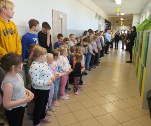 Grupa uczniów różnych klas stoi w rzędzie na szkolnym korytarzu podczas uroczystości, z przypiętymi żółtymi żonkilami.