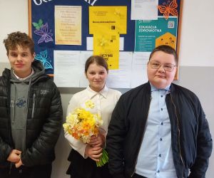 Trójka uczniów stoi przed tablicą ogłoszeń samorządu uczniowskiego, trzymając bukiet żółtych żonkili.