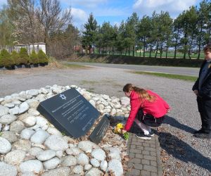 Uczennica składa bukiet żółtych żonkili przy tablicy upamiętniającej ofiary, pozostali uczniowie stoją obok.