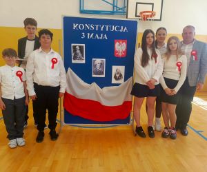 Uczniowie uczestniczący w apelu wraz z nauczycielem przygotowującym uroczystość na tle tablicy dekoracyjnej