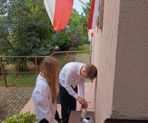 Uczennica i uczeń składają kwiaty i zapalają znicz pod tablicą pamiątkową patrona szkoły