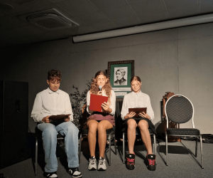 Troje uczniów siedzi na scenie podczas prezentacji fragmentów „Dziadów”. Jedna z uczennic czyta tekst do mikrofonu, a pozostali śledzą tekst w czerwonych teczkach. W tle znajduje się portret Adama Mickiewicza.