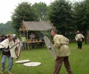 Dwóch mężczyzn w strojach antycznych z tarczami i mieczami, w tle stoisko z artykułami z gliny 