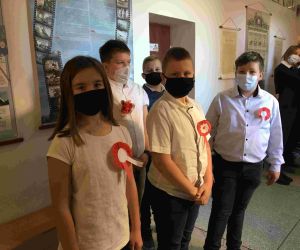 Uczniowie klasy V w strojach galowych i maskach, z przypiętymi kotylionami w barwach narodowych.