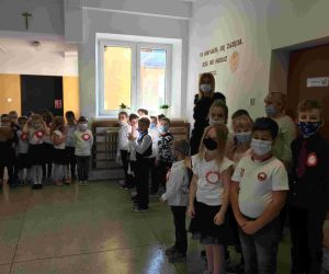 Grupy uczniów w strojach galowych i maskach z panią dyrektor szkoły na korytarzu głównym. Wszyscy mają przypięte kotyliony w barwach narodowych.