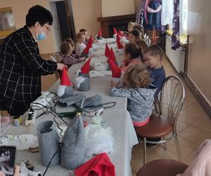 Dzieci siedzą przy stole, coś robią. Dorosły pokazuje im czynności, jakie muszą wykonać.