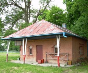 Budynek z czerwonej cegły, na przodzie trzy kolumny podpierające dach wykonane z drzew pomalowanych na biało-czerwono, w tle drzewa i krzewy