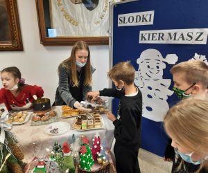 Uczennica za stołem wręcza kawałek ciasta uczniowi