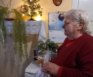 Starsza kobieta trzyma stroik świąteczny w dłoni. W tle wnętrze pokoju z małą oświetloną choinką