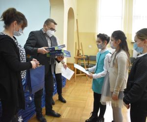 Dzieci i osoby dorosłe wewnątrz budynku. Mężczyzna wręcza nagrody dyplom.