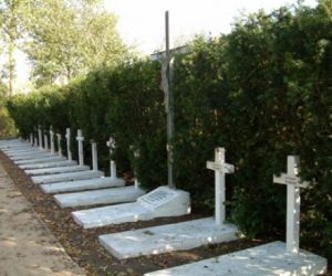 Białe murowane mogiły z murowanymi krzyżami, na jednej duży metalowy krzyż, za nimi wysoki żywopłot