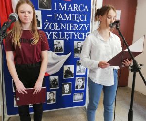 Dwie uczennice przy mikrofonach. W tle tablica poświęcona Żołnierzom Wyklętym