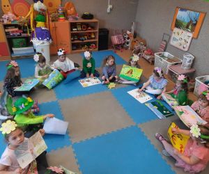 Przedszkolaki w strojach wiosennych siedzą w kręgu. Trzymają w rękach prace plastyczne o tematyce wiosennej