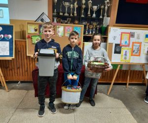 Uczniowie stoją na korytarzu,  zamiast plecaków mają śmietniczkę, wiadro i wiklinowy koszyk.