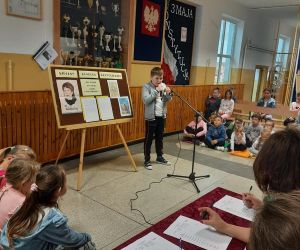 Uczeń ubrany w szarą bluzę recytuje wiersz. Uczniowie siedzą obok i słuchają go, jury ocenia recytację.