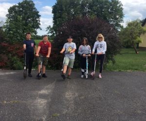 Uczniowie z nauczycielem – każdy z hulajnogą – stoją na tle zieleni przed szkołą