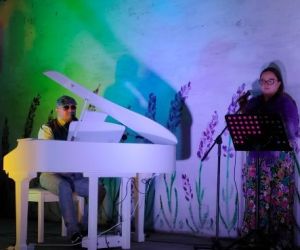 Osoba gra na fortepianie wewnątrz budynku, a druga osoba śpiewa.