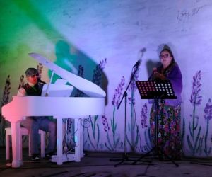 Osoba gra na fortepianie wewnątrz budynku, a druga osoba śpiewa.