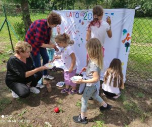 Na zdjęciu widoczna jest grupa dzieci malująca wraz z paniami na prześcieradle wywieszonym na płocie przed budynkiem.