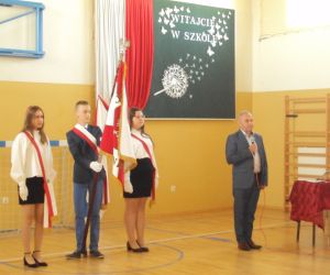 Pan dyrektor wygłasza przemowę.  W tle poczet sztandarowy – chłopiec trzyma sztandar obok dwie dziewczynki.