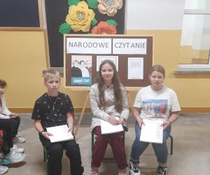 Uczniowie siedzą na krzesełkach i czytają Ballady A. Mickiewicza, za nimi stoi tablica z informacjami z okazji Narodowego Czytania 2022.