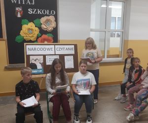 Nauczycielka czyta list Prezydenta RP z okazji 11 akcji Narodowego Czytania, uczniowie siedzą na krzesełkach i wsłuchują się w ten list.