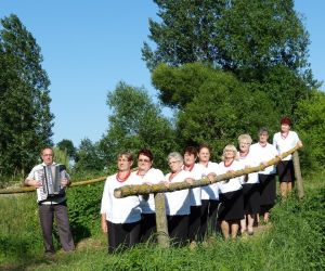 Członkinie zespołu śpiewaczego i Klubu Seniora BIEDRONKI z instruktorem. Ubrani uroczyście, pozują do zdjęcia w otoczeniu natury.