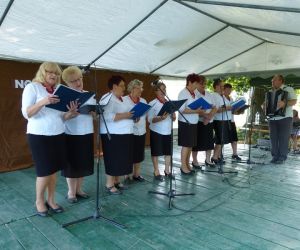 Kobiety stojące na scenie. Przed nimi mikrofony. W rękach trzymają teczki. Przed ni mi mężczyzna z akordeonem. 