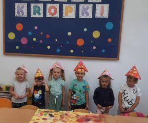 Przedszkolaki w ubraniach i papierowych czapeczkach w kropki na tle tablicy dekoracyjnej