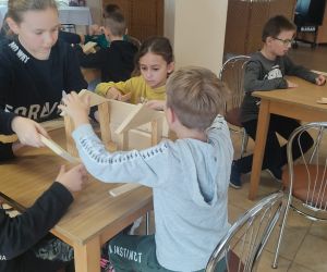 Na zdjęciu są widoczne dzieci siedzące przy stolikach składające elementy karmnika dla ptaków.