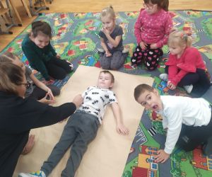 Nauczyciel odrysowuje postać chłopca na szarym papierze, a dzieci się przyglądają