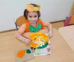 Dziewczynka przy stoliku maluje farbami dynię na papierowym talerzyku